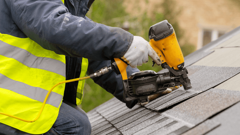 Technician installing shingles on a roof while wearing yellow safety vest.