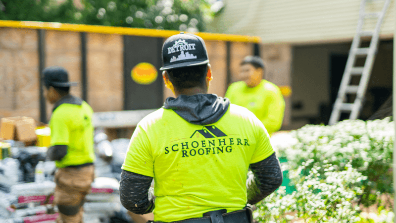 Team of roofers, one in foreground with back to camera, all wearing yellow t-shirts that say "Schoenherr Roofing" on the back. Standing outside working.